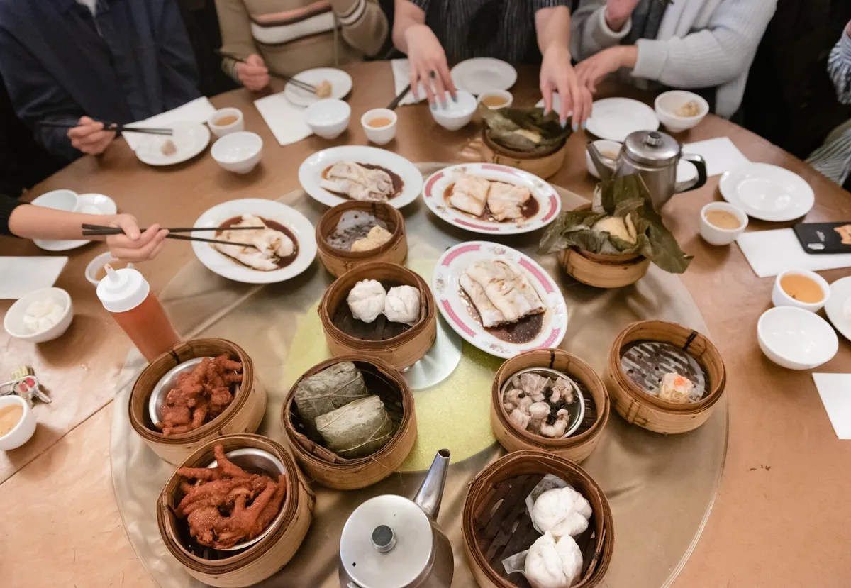 A top-down photograph of a number of steamers and small plates of dim sum atop a lazy susan turntable. Hands holding chopsticks and reaching for dim sum reach from out of frame.